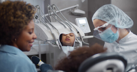 African little girl sitting in dental chair and looking in mirror visiting dentist with mother
