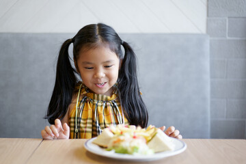 Asian child hungry or kid girl smile to enjoy happy eating bread or sandwich and vegetable salad on dish and wood table at school or home kitchen and restaurant for breakfast food or snack at lunch