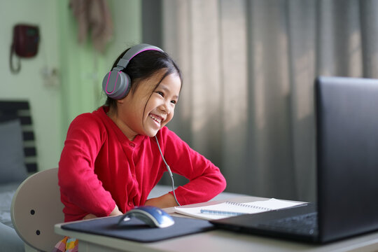 Asian child or kid girl smile enjoy e-learning on computer notebook with wearing headphone to live streaming communication and study online or people learn from home to play laptop by back to school