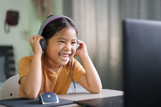 Asian Child Student Or Kid Girl Smile Enjoy E-learning On Computer Notebook With Wearing Headphone For Laugh To Funny Communication And Study Online Or Learn From Home To Play Laptop By Back To School