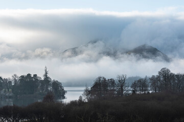 Dramatic landscape image looking across Derwentwater in Lake District towards Catbells snowcapped mountain with thick fog rolling through valley