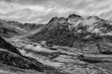 Obraz premium Stunning flying drone black and white landscape image of Langdale pikes and valley in Winter with dramatic low level clouds and mist swirling around