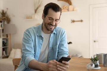 Happy young Caucasian man sit at desk at home look at smartphone screen texting or messaging online on gadget. Smiling millennial male use cellphone, browse wireless internet. Mobile provider concept.