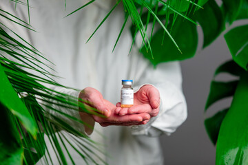 Woman in white shirt show vaccine in a bottle near palm leaves.