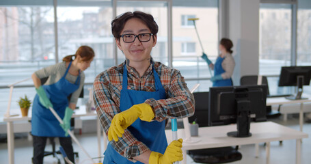 Asian janitor in glasses and uniform smiling at camera with colleagues working on background