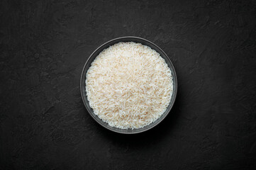 Raw long grain rice in black ceramic bowl on dark slate table top. Heap of uncooked basmati or jasmine rice. Healthy vegetarian organic food ingredient