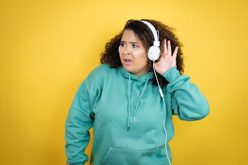 Young african american girl wearing gym clothes and using headphones surprised with hand over ear listening an hearing to rumor or gossip