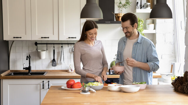 Wide banner panoramic view of happy millennial Caucasian couple cooking healthy tasty vegetarian salad in home kitchen together. Smiling young man and woman prepare delicious diet food for breakfast.
