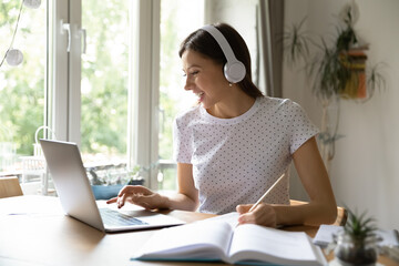 Happy millennial Caucasian female student in headphones look at laptop screen study distant from home. Smiling young woman in earphones make notes watch webinar on computer, take web course.