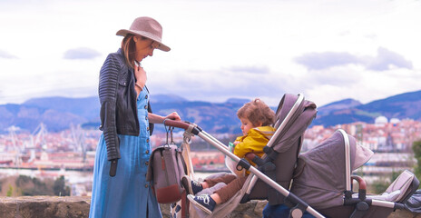 Beautiful middle aged young woman leaning wearing leather jacket hat and blue dress pushing a twin...