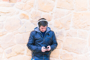 Attractive young middle-aged man leaning on stone wall wearing wireless music headphones and leather jacket looking at smartphone