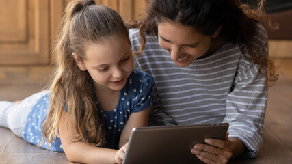 Close up smiling young mother and little daughter using tablet together, lying on warm floor with underfloor heating, family having fun with gadget, browsing apps, watching video, playing games