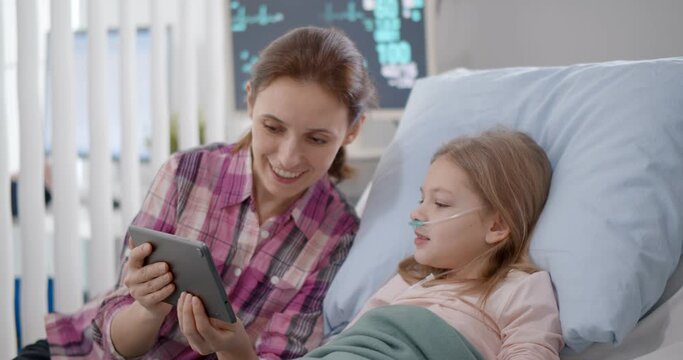 Mother and daughter in hospital bed watching cartoons on digital tablet