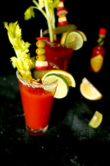 Two glasses of Bloody Mary cocktail garnishes with celery, olives, pickles, cheese and lime, black background