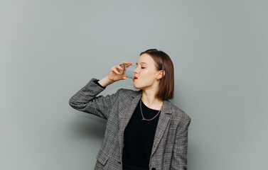 Beautiful female office worker in a jacket and formal clothes drinks water from a glass on a background of gray wall. Business lady quenches thirst with a glass of water. Isolated.