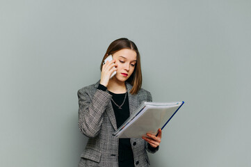 Fototapeta premium Young female office worker in smart casual clothes stands on a gray background and calls on a smartphone, reads documents with a serious face. Lady is in business, isolated.