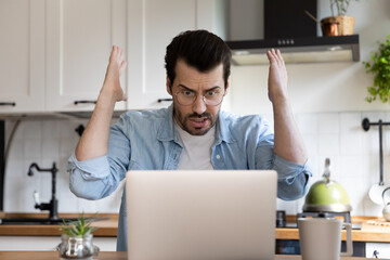 Unhappy mad millennial man look at laptop screen shocked by online virus or spam on web on gadget....