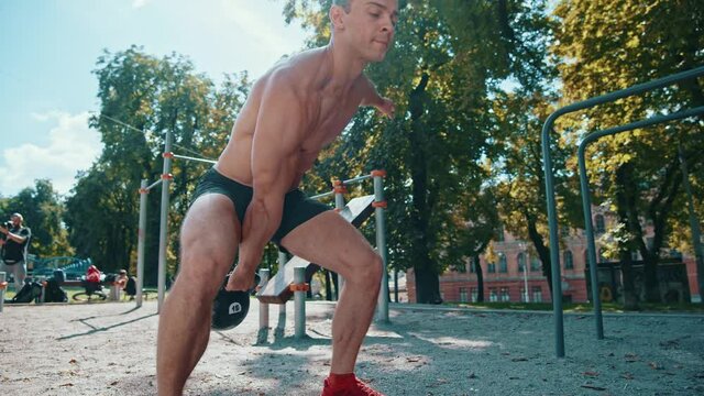 Low angle view of the strong athletic man training with weight at the street. Caucasian guy throwing up a sports kettlebell while exercising at the sunny summer day. Sport and recreation concept