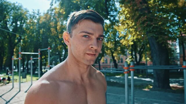 Portrait view of the strong guy training outdoors. Serious caucasian man looking to the camera with calm emotions. Handsome man posing with horizontal bars at background. Sport and training together