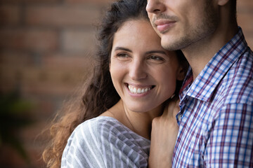 Fototapeta premium Close up smiling beautiful dreamy woman hugging man, looking to aside, happy young wife visualizing good future with husband, young couple enjoying tender moment, planning, standing at home