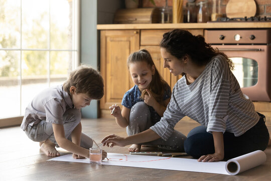 Caring mother and little kids painting colorful watercolors at home together, sitting on warm wooden floor in kitchen, happy young mum with adorable preschool son and daughter enjoying leisure time