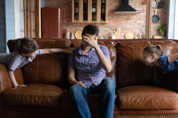 Exhausted unhappy father having problem with noisy naughty kids, sitting on couch, touching...