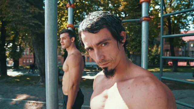 Portrait of strong guy training outdoors. Serious caucasian unshaven man training with friend. Man posing with horizontal bars at background. Sport and training together