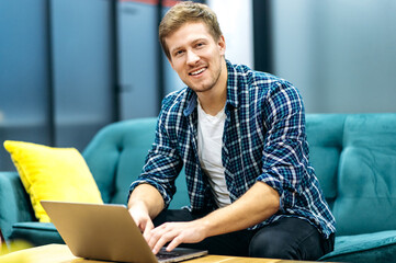 Handsome caucasian guy freelancer uses laptop. Smiling attractive guy in stylish casual sits on a sofa in a creative office, browsing internet, looks and smiles friendly at the camera