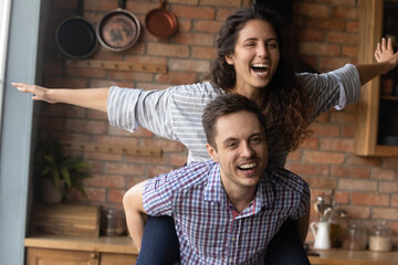 Close up smiling husband carrying excited wife pretending flying with hands outstretched, young...