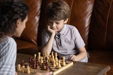 Close up thoughtful little boy playing chess with mother, pondering strategy, family enjoying...