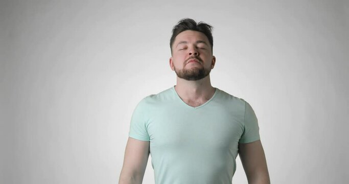 Young bearded man in basic green t-shirt takes off medical mask and makes a beed breath on grey background, front view. Covid-19, coronavirus pandemic, epidemic, quarantine. Protection from virus.