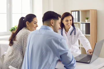 Smiling nurse showing ultrasound baby scan picture to future parents. Couple of happy young patients, husband and his pregnant wife, looking at computer screen sitting at table at doctor's office