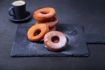 Donuts with powdered sugar and a cup of coffee. Traditional donuts in the shape of a ring fried in oil, on a black background. Junk food. Close-up.