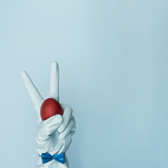 Gloved hand holds an Easter red egg on a pastel, trendy blue background. Dressy rabbit with copy space. Minimal Easter concept. Corona virus style.