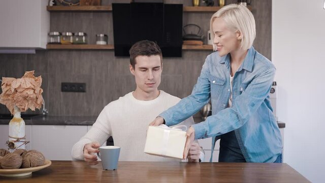 Young beautiful couple at home in the kitchen. A blonde woman gives her husband a gift for a holiday.