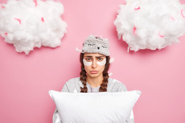 Tender cute young European girl with two combed pigtails poses in nightwear applies beauty patches holds soft pillow wears sleeping eyemask looks sadly at camera isolated over pink background
