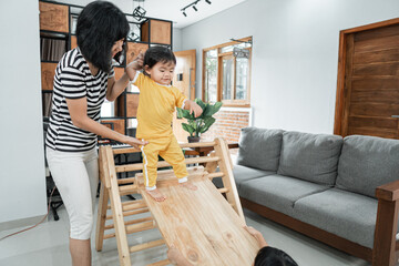 a mother patiently holds the baby's hand while standing sliding on a pikler triangle toy at home