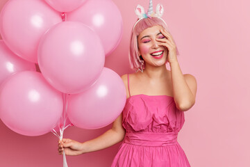 Positive Asian woman with pink hair giggles positively keeps hand on face has bright professional makeup wears dress has fun at party holds inflated helium balloons isolated over pink background