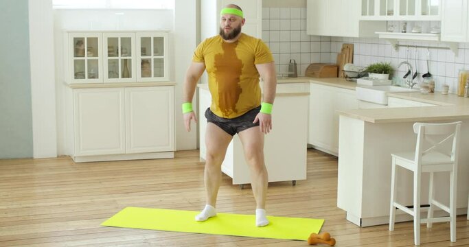 Bearded fat sweaty man newcomer in sport in yellow wet sportswear doing squats exercise training at home on fitness mat. Sport, workout, losing weight concept. Joke, mem, fun behavior, humor.