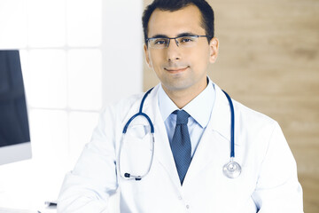 Man-doctor sitting at the desk at his working place and smiling at camera. Perfect medical service in clinic