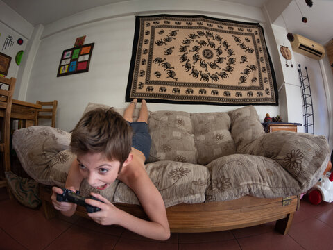 Ni&ntilde;o juega con joystick divertido tirado en el sof&aacute; a punto de caerse
