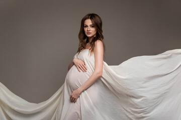 Pregnant girl in white flying cloth. Keeps belly. Grey background.