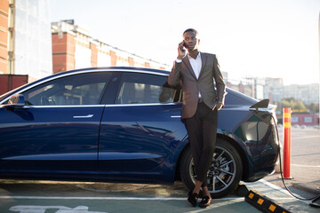 Fototapeta na wymiar Electric car charging concept. Full length portrait of young handsome dark skinned man, standing at the charging station and talking phone whie waiting for his car charging