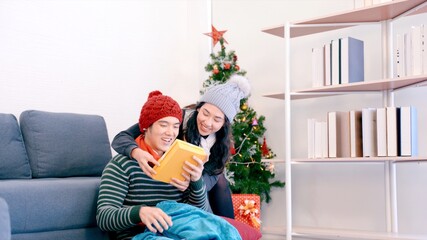 Obraz na płótnie Canvas Asian young is getting an amazing Gift from his Wife. Young Couple is celebrating Christmas, New Year at Home, Cozy Festive Evening. Happy holiday in winter time.