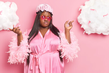 Horizontal shot of serious overweight Afro American lady with long dark hair indicates aside shows something wears bath hat silk dressing gown isolated over pink background white clouds above