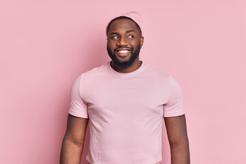 Cheerful bearded black man smiles broady looks curiously aside has white even teeth wears hat and t shirt in one tone with background. Pleased Afro American guy notices something interesting