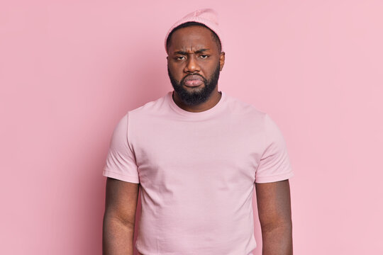 Photo of frustrated displeased bearded dark skinned man looks unhappily at camera being dissatisfied with something wears hat and casual t shirt isolated over rosy background. Negative emotions