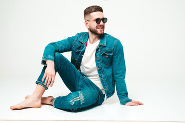 Portrait of handsome smiling stylish hipster lambersexual model.Man dressed in jacket and jeans clothes. Fashion male sitting on grey background in studio in sunglasses