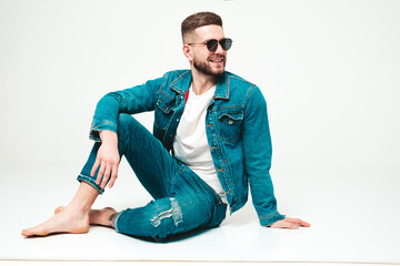 Fototapeta premium Portrait of handsome smiling stylish hipster lambersexual model.Man dressed in jacket and jeans clothes. Fashion male sitting on grey background in studio in sunglasses