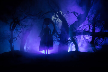 Fototapeta premium Horror Halloween decorated conceptual image. Alone girl with the light in the forest at night. Silhouette of girl standing between trees with surreal light on background. Selective focus.